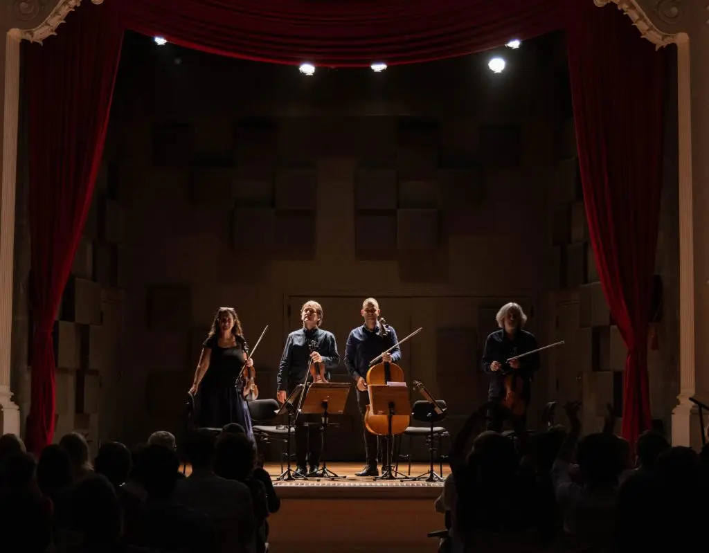 Quatro músicos se apresentam no palco de um teatro diante do público, com instrumentos de cordas, incluindo violino, violoncelo e violão. O palco é emoldurado por cortinas vermelhas e iluminado por refletores.