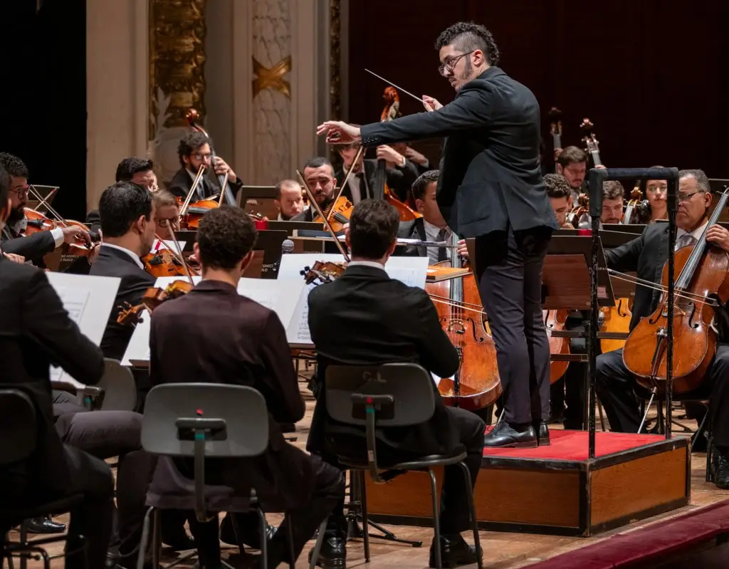 Maestro rege uma orquestra sinfônica durante uma apresentação em um teatro. Músicos sentados tocam instrumentos de cordas, como violinos e violoncelos, enquanto acompanham a regência no palco.