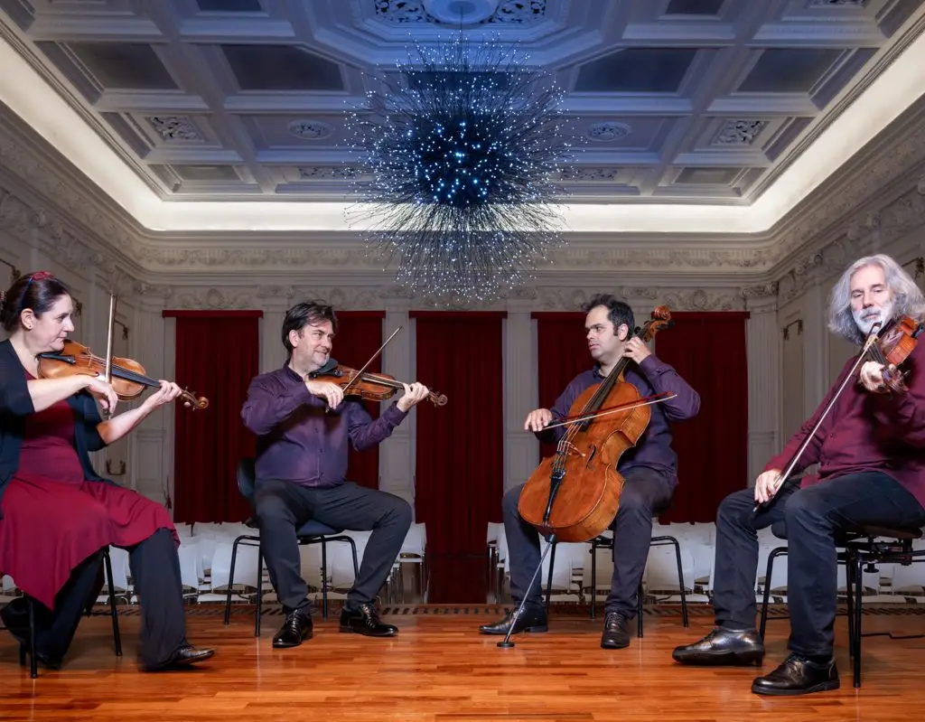Quatro músicos sentados tocam instrumentos de cordas em um elegante salão ornamentado, com cortinas vermelhas, piso de madeira e um grande lustre iluminado em azul no teto.