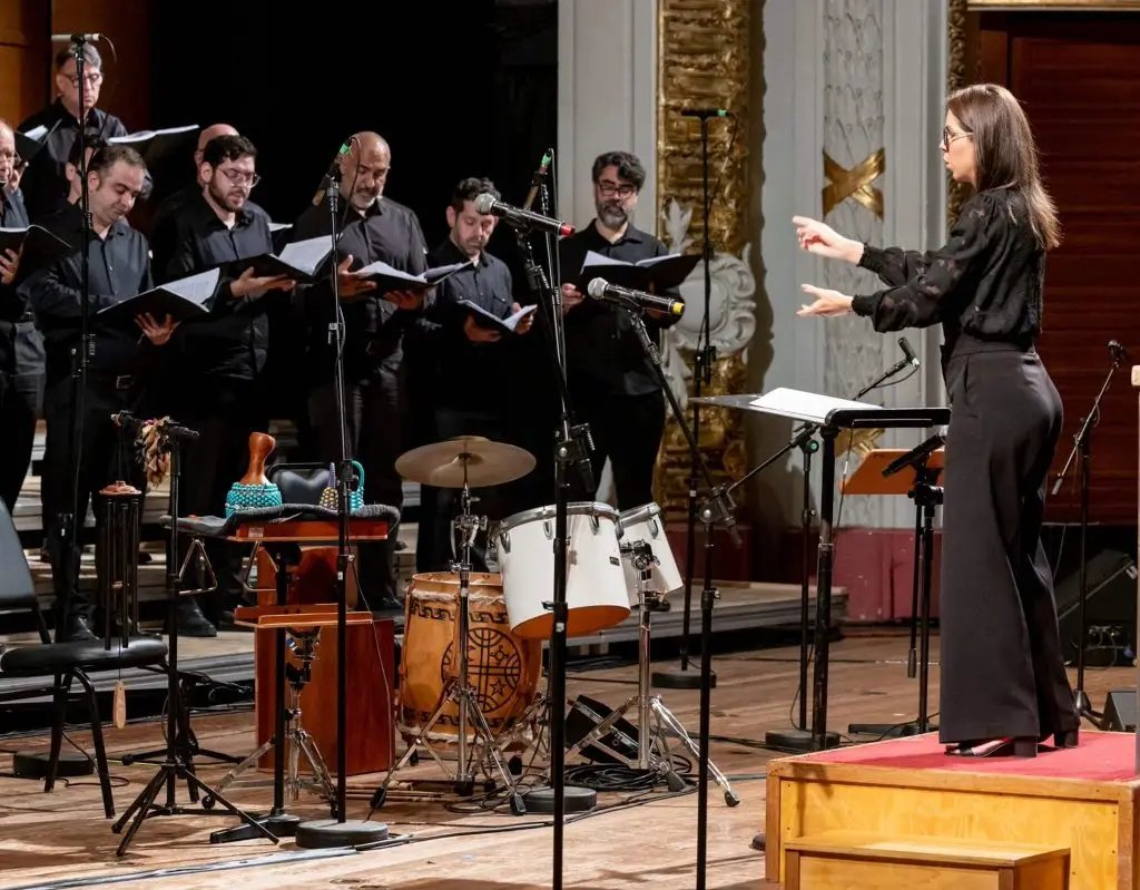 Uma mulher conduz um grupo de cantores durante uma apresentação musical em uma igreja. Os músicos, vestidos de preto, seguram partituras e estão posicionados ao lado de diversos instrumentos de percussão, microfones e estantes.
